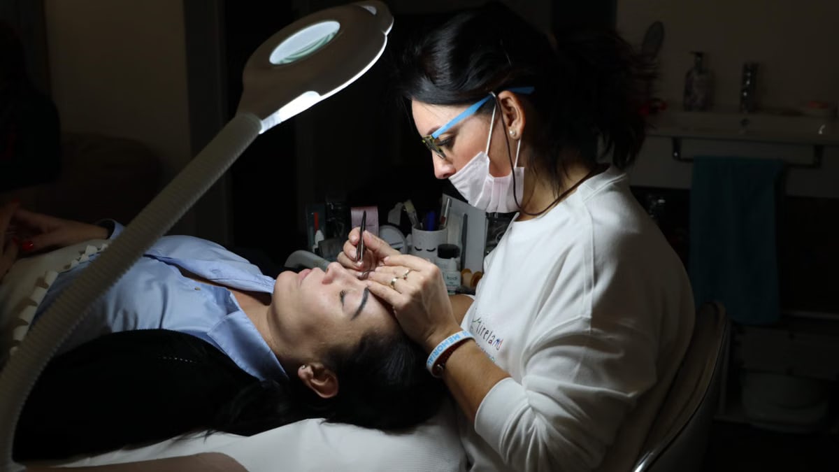 A beautician provides professional eyebrow care to a client in a salon, under focused lighting.